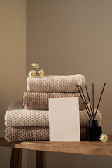 Stack of towels in neutral colors, aroma diffuser and paper blank in modern aesthetic interior. Fabric bathroom accessories for hygiene, spa and skin care.