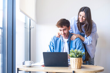 Happy young couple sitting at home together, smiling while looking at laptop and documents....