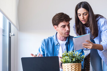 Young couple sitting at home reviewing documents and laptop together, focused on managing household expenses, bills, or investments. Serious expressions reflect financial planning, budgeting, and life