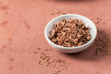A white bowl filled with chocolate shavings sits on a reddish-brown surface, with scattered...