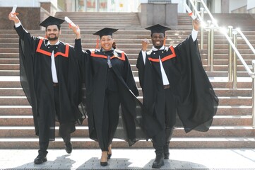 Group of happy smiling multicultural people in graduation gowns and caps with diplomas in hands...