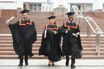Group of happy smiling multicultural people in graduation gowns and caps with diplomas in hands...