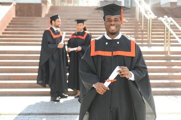 Happy graduate. Happy African man in graduation gowns holding diploma and smiling while his friends...