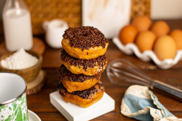 Donat Kampung or Donut Chocolate Sprinkles stacking, concept of chocolate doughnuts served with white square coaster, mug, blue napkin, flour on mini wooden bowl, bamboo tray, eggs, whisk and milk.