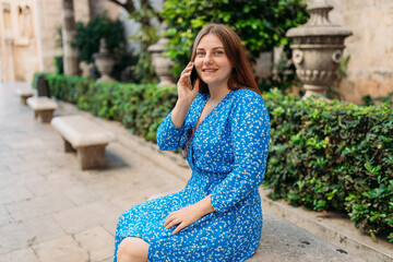 Young happy redhead 30s woman making phone call over mobile phone in the city. Middle age woman talking on smartphone on urban background. Traveling Europe in summer