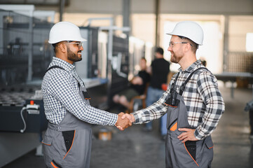 Two engineers in hard hats shake hands in a factory.