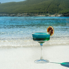 A clear, blue-toned cocktail, with a dried blood orange slice on the rim of the glass as a...