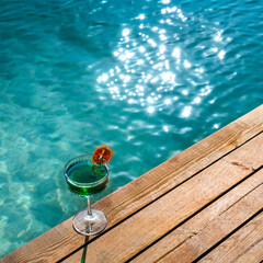 A refreshing, clear, blue-toned summer drink cocktail placed on a wooden pier by the beach, with a...