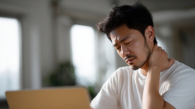 Young Japanese man experiencing neck pain while working on laptop, showing discomfort and stress in modern workspace. scene captures challenges of remote work and physical strain