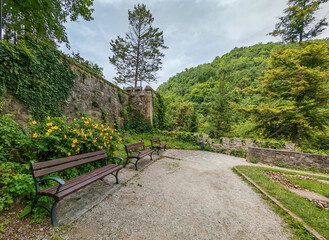 Wooden benches line a peaceful corner of the Lillafüred Hanging Garden, surrounded by blooming yellow flowers and an ivy-covered stone wall, with lush green hills in the background.