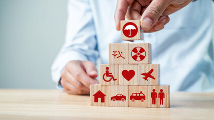 Hand placing a wooden block with an umbrella symbol on a stack of blocks representing various life risks and assets for insurance coverage
