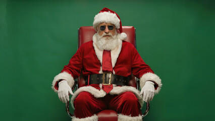 Cool santa claus wearing sunglasses and a red suit sits confidently in a chair against a green background