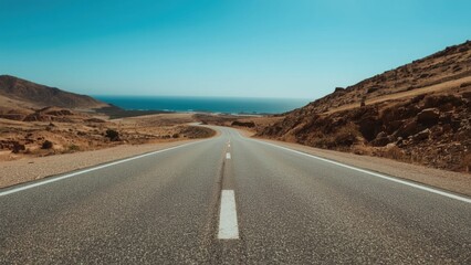 Empty asphalt road stretching through arid desert landscape towards the horizon with a view of the sea under a clear blue sky