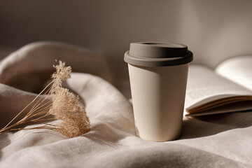 Reusable coffee cup on linen tablecloth with book and dried flower