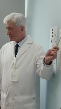 Vertical shot of senior male eye doctor dressed in white coat pointing at chart with Landolt rings examining patients eyesight during visual acuity test in hospital
