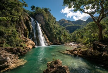 Obraz premium Majestic waterfall cascading into a serene turquoise pool surrounded by lush greenery and tropical mountains under a clear blue sky in nature setting