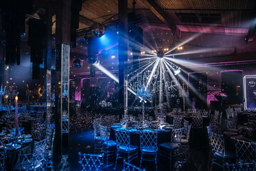 Description: Luxurious event space featuring round tables set for dining with elegant tableware and crystal chairs. Ambient lighting enhances the sophisticated atmosphere for gatherings or celebration