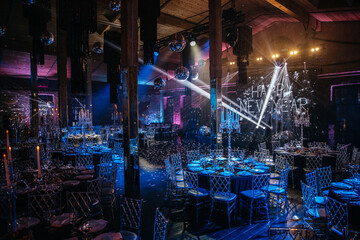 Description: Luxurious event space featuring round tables set for dining with elegant tableware and crystal chairs. Ambient lighting enhances the sophisticated atmosphere for gatherings or celebration