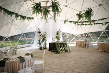 Elegant setup for a wedding reception inside a geodesic dome, featuring beautifully set tables with floral decorations and greenery. Minimalist design creates a serene atmosphere.