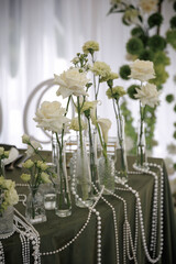 Elegant table setting featuring tall white roses and delicate flowers in glass vases, intertwined with pearl strands on a dark cloth, creating a sophisticated atmosphere.