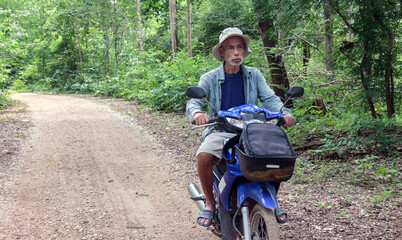 An elderly Asian man rides a motorbike on a dirt road in the forest.