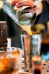 Bartender pouring a yellow cocktail through a strainer into a metal cup. Close-up of professional...