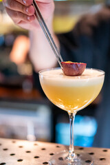 Bartender garnishing elegant yellow cocktail with halved passion fruit in coupe glass. Close-up of stylish drink preparation at bar counter.