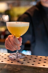 Cocktail glass with yellow drink and foam held by bartender's hand, set against a blurred bar...