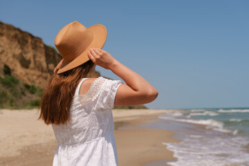 Close-up of a woman in a white dress and hat holding her hat while looking at the ocean on a sunny...