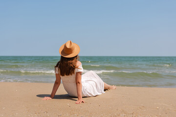 Woman in white dress and hat sits on sandy beach, facing the sea and enjoying the peaceful ocean...