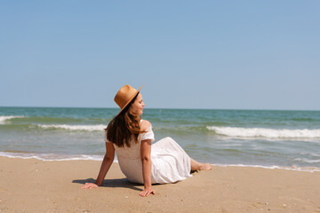 Young woman in white dress and hat relaxes on sandy beach, enjoying the sea breeze and sunshine...