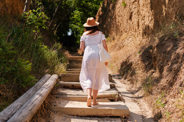 Barefoot woman in a white summer dress and hat walking up rustic forest steps, surrounded by dry cliffs and greenery, on a sunny day with a canvas bag in hand.