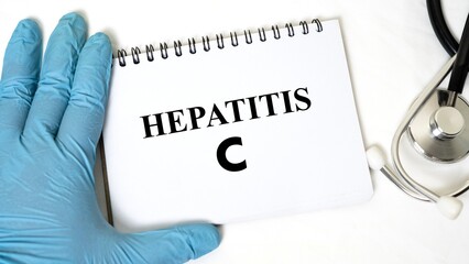 Close-up image of a gloved hand holding a spiral notebook with the word "HEPATITIS C" written on it, accompanied by a stethoscope on a white background.