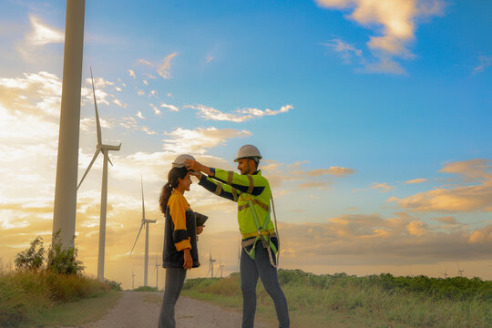 Male engineer gives safety helmet attentive efficiency to female engineer for safety, accident prevention and professionalism during field work at wind turbine station : Safe working concept.