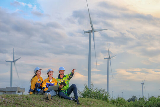 Team male and female engineers sit together grassy hill at wind farm sharing concept about renewable energy from the earth and reviewing plans as wind turbines stand tall against cloudy sunset sky.