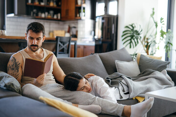 Couple relaxing at home, man reading a book while his girlfriend is sleeping on his lap, resting on the sofa in the living room