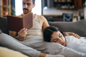 Couple relaxing on the sofa at home, he is reading a book while she sleeps peacefully on his legs