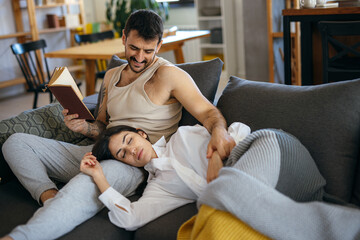 Couple relaxing together on the sofa at home, with the man engrossed in a book while the woman peacefully sleeping on his lap
