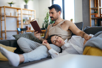 Couple relaxing on the sofa at home, the man is reading a book while his girlfriend is sleeping with her head on his lap