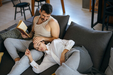 Young man reading a book and caressing his girlfriend sleeping on his lap on a comfortable sofa at home