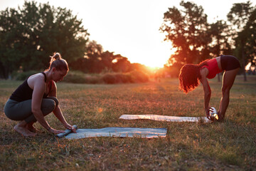 Two women folding and unfolding yoga mat in the park during sunset sunrise time.