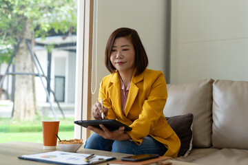 Asian woman yellow blazer digital tablet stylus working living room relaxed morning
