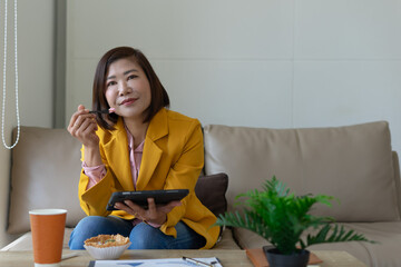Woman in yellow blazer eating dessert, holding tablet, sitting on sofa, relaxed and thoughtful