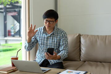 Man glasses plaid shirt sitting sofa laptop smartphone video call waving relaxed