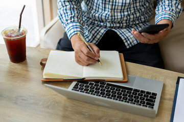 Man writing in notebook with pen while holding smartphone, laptop and iced coffee on wooden table