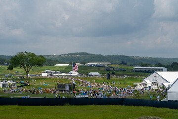 Obraz premium A large golf course is filled with spectators and event tents under a cloudy sky. An American flag stands prominently amidst the crowd, with distant hills in the background.