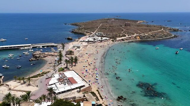 Drone view of bustling beach with many swimmers and sunbathers curves around a clear turquoise bay