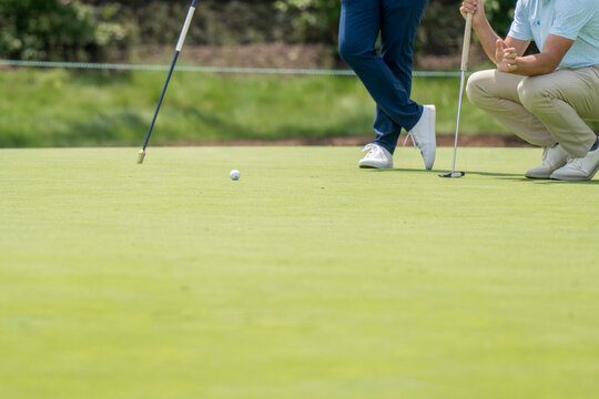 Two golfers are on a vibrant green putting surface; one stands with a putter, while the other crouches to observe a golf ball, indicating focus on precision in the game.
