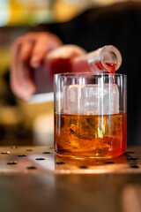Close-up of a hand pouring red liquid from a bottle into a glass with ice cubes and amber drink, on...
