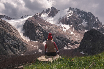 Rear view of a woman sitting cross-legged on a rock, wearing a red beanie and vest, meditating or resting while facing dramatic snow-covered mountain peaks under a cloudy sky. 